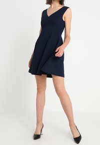 Tmavě modré bezramínkové šaty s výstřihem do V, rozšířenou sukní a plisováním. Materiál vypadá hladce a má strukturovaný střih. Nosené s černými podpatky.