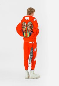 Sudadera naranja con un diseño de alas multicolores en la parte posterior, combinada con pantalones jogger a juego que tienen acentos blancos. Botas blancas completan el conjunto.