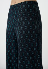 Zwarte gestreepte broek met een geometrisch patroon van teal pijlen, gemaakt van een zachte stof met een gladde textuur.