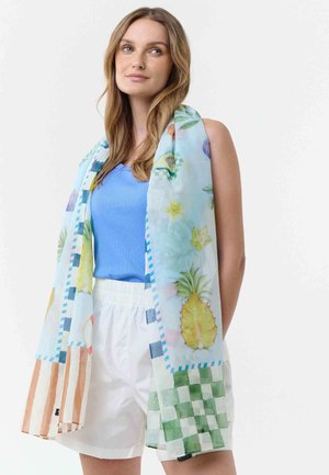 Frau trägt ein blaues Oberteil und weiße Shorts, die mit einem leichten Schal mit tropischen Früchten und karierten Mustern drapiert sind.