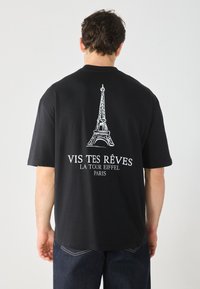 Black T-shirt with white Eiffel Tower graphic and text "VIS TES RÊVES LA TOUR EIFFEL PARIS" on back; short sleeves, relaxed fit.
