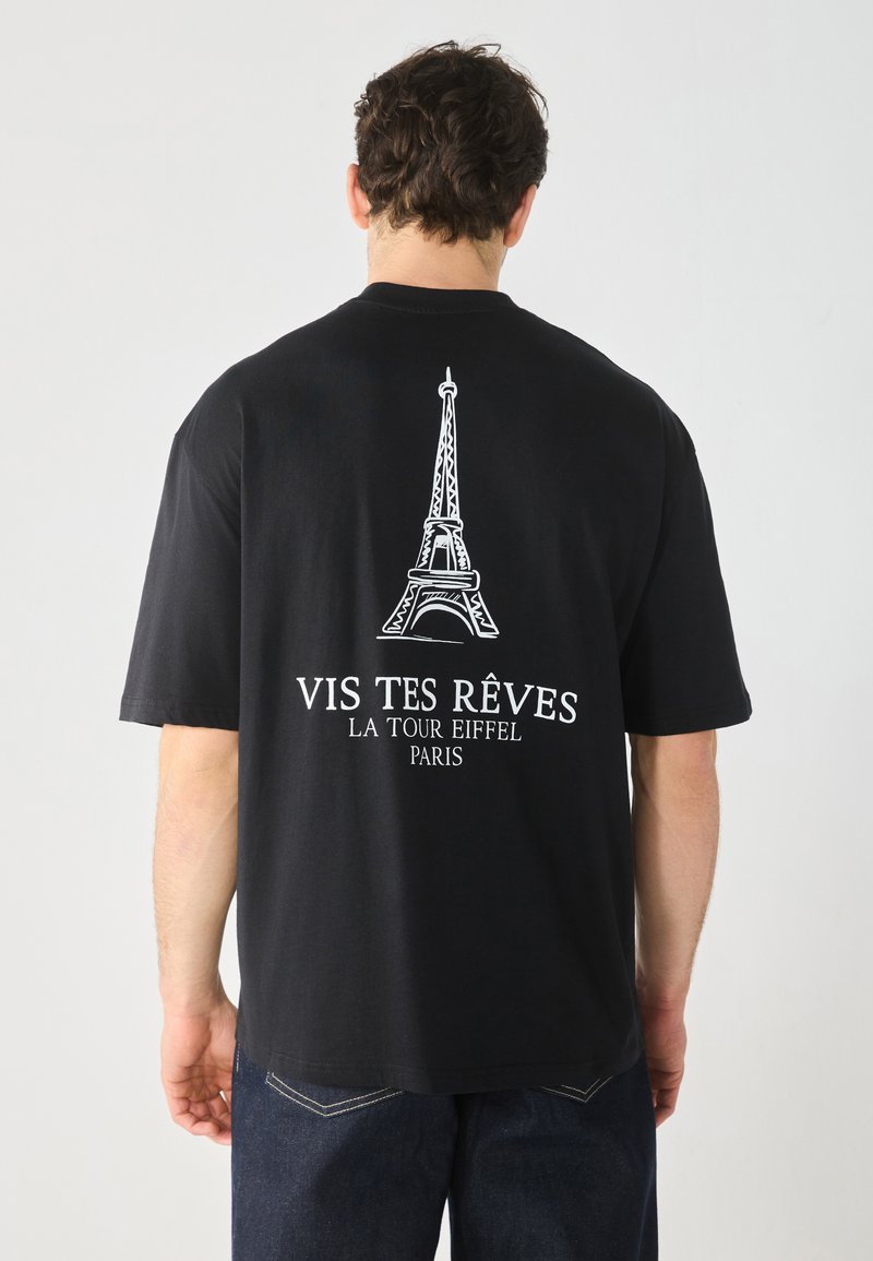 Svart T-skjorte med hvit Eiffel-tårn grafikk og teksten "VIS TES RÊVES LA TOUR EIFFEL PARIS" på baksiden; korte ermer, avslappet passform.