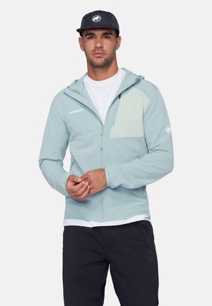 Mann trägt hellblaue Jacke mit Reißverschluss und Kapuze, weißes T-Shirt, schwarze Kappe und schwarze Hose, steht vor einfachem Hintergrund.