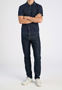 Granatowa dzianinowa koszulka polo z krótkim rękawem ozdobiona fakturowanym wzorem warkocza, kołnierzyk zapinany na guziki, zestawiona z ciemnymi dżinsami i białymi trampkami.