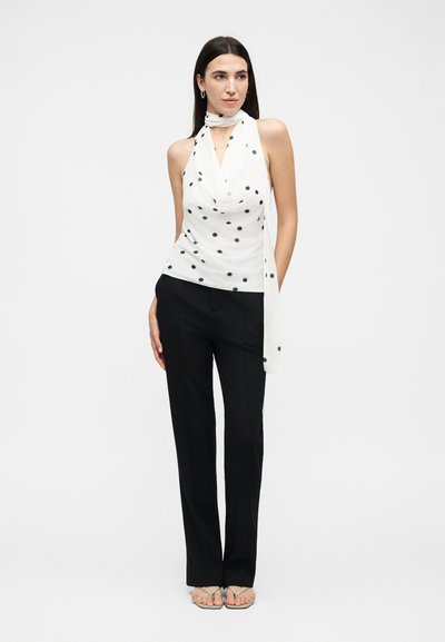 Femme portant un haut blanc sans manches à col halter avec des pois noirs, un pantalon noir droit et des sandales beiges sur fond blanc.