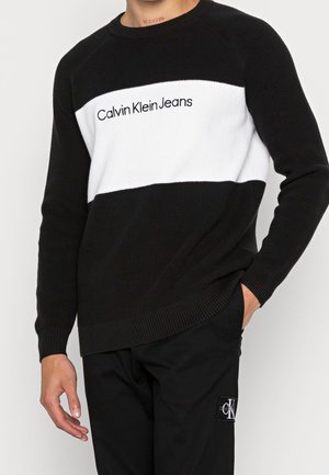 Mann trägt einen schwarz-weißen Calvin Klein Jeans Pullover und schwarze Hosen mit Calvin Klein Logo-Aufnäher an der Tasche, Hand in der Tasche.