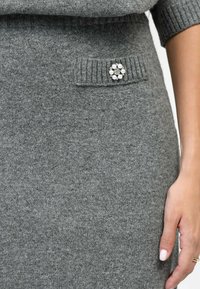 Jupe en tricot gris avec une poche côtelée ornée d'un broche décorative en perles circulaires. Texture lisse avec une silhouette ajustée.