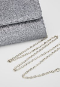 Silverglittrande kuvertväska med fälllock och avtagbar silverkedja samt en texturerad, glittrande yta.