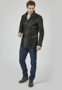 Giubbotto in pelle nera con colletto rigido, chiusura frontale con zip e molte tasche. Abbinato a jeans blu e stivali marroni con lacci.