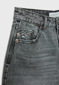 Zbliżenie na kieszeń szarych dżinsów z denimu z nitami, pętlą na pasek i małą metalową metką z logo na kieszonce na drobne.