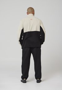 Giacca in pile beige e nera, con collo alto, logo sul retro, abbinata a pantaloni neri coordinati.