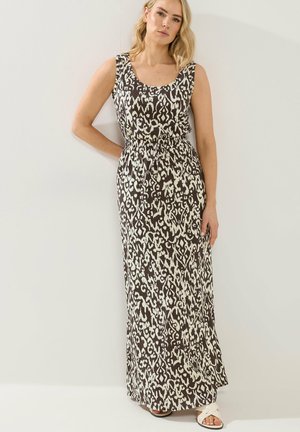 Frau mit blondem Haar, die ein ärmelloses Maxi-Kleid mit schwarz-weißem abstraktem Muster und weiße Sandalen trägt, steht vor einer schlichten Wand.