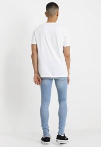 Mies seisoo selkä kameraan päin, päällään yksinkertainen valkoinen T-paita, vaaleansiniset tiukat farkut ja mustat lenkkarit, joissa on valkoiset pohjat.