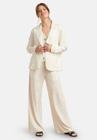 Beige Nadelstreifenanzug mit einem einreihigen Blazer mit zwei Knöpfen und weit geschnittenen Hosen, kombiniert mit schwarzen Schuhen.