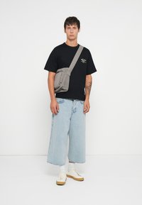 Borsa a tracolla grigia in tessuto, indossata su una maglietta nera e jeans a gamba larga blu chiaro. Presenta una tasca frontale con zip e una tracolla regolabile.
