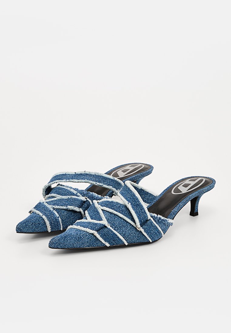 Un paio di mules in denim blu a punta, con bordi sfrangiati, design a cinturini incrociati e tacco kitten basso su uno sfondo bianco.