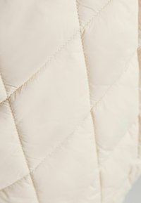 Světle béžová prošívaná bunda s kameninovým vzorem, hladkou texturou a jemným leskem. Podél švů jsou viditelné prošívané detaily.