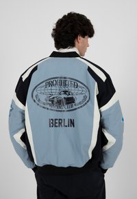 Homme portant une veste bleue et noire avec un graphique de voiture de course et le texte "Prohibited Racing Club Berlin" au dos, tourné vers l'arrière.