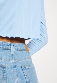 Vaaleansininen ribbineulottu lyhyt toppi siksakreunuksella ja pitkillä hihoilla, yhdistetty korkeavyötäröisiin vaaleisiin farkkuihin, joissa on nahkainen merkintätasku.