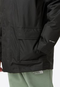 Černá voděodolná bunda s texturovaným povrchem, se skříňovou kapsou a logem na rukávu. Spárována s světle zelenými kalhotami.