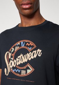 Juodas medvilnės marškinėlis su dideliu, daugiakampiu "Sportswear" grafiniu piešiniu ir stilizuotomis raidėmis. Trumpomis rankovėmis, apvalia iškirpte.