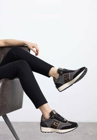 Zapatillas deportivas negras y beige con detalles en estampado de leopardo, tela texturizada y una suela blanca gruesa. Leggings negros complementan el calzado.