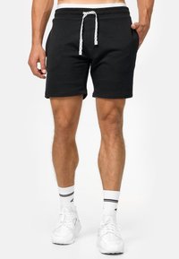 Sorte bomulds shorts med et tætsiddende design, hvid snor, sidelommer og stikninger i kanten. Båret med hvide sneakers og stribede sokker.