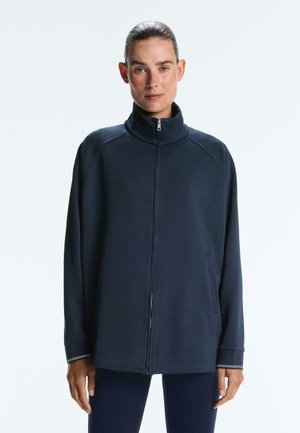 Žena s vlasmi zviazanými dozadu, oblečená v námornícky modrej bunde na zips s vysokým golierom a dlhými rukávmi na svetlom pozadí.