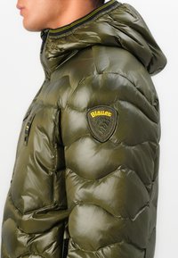 Veste matelassée vert olive avec capuche, présentant une texture brillante, un design matelassé et un patch logo jaune et noir sur l'épaule.