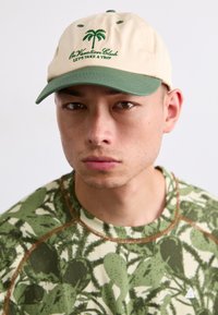 Baseballcap i creme med grøn skygge, der har broderede palmetræer og tekst. Båren med en grøn mønstret skjorte med en tekstureret finish.