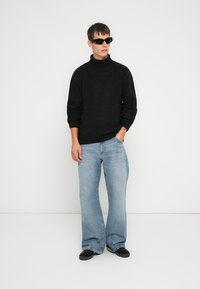 Svart høyhalset genser med teksturert strikk, i kombinasjon med lyseblå vide jeans. Modellen har på seg kantete svarte solbriller og svarte sko.