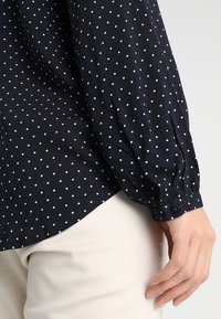 Námořnická modrá blůza s bílými puntíky, splývavá látka, volné rukávy a knoflíkový detail na manžetě, doplněná o světlé kalhoty.