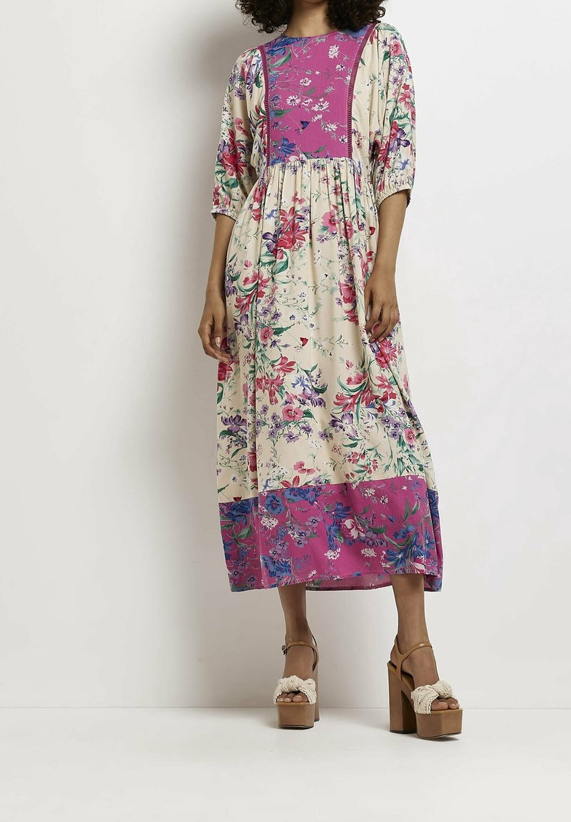 Femme portant une robe midi fleurie crème avec des panneaux roses, des manches bouffantes et des sandales à plateforme marron, debout sur un fond blanc.