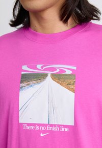 Lyserød bomulds T-shirt med et grafik af en vej, med svungne design ovenfor og teksten "Der er ingen mållinje" i hvidt, med Nike-logoet nedenfor.