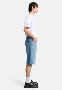 Shorts en denim bleu clair à coupe droite, avec des poches à l'avant et un ourlet légèrement effiloché. Associés à un t-shirt blanc et des chaussures noires.