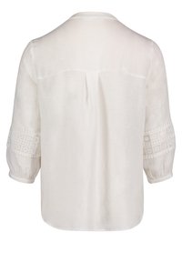 Blusa bianca con una vestibilità ampia, caratterizzata da decorazioni in pizzo sulle maniche e una piega nella parte posteriore. Tessuto liscio con una texture leggera.