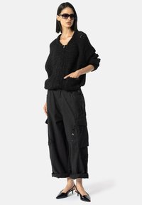 Pulover negru tricotat, cu decolteu în V și croială oversized, asortat cu pantaloni cargo negri, ornamentați cu broderie florală și manșete răsucite. Tocuri negre.