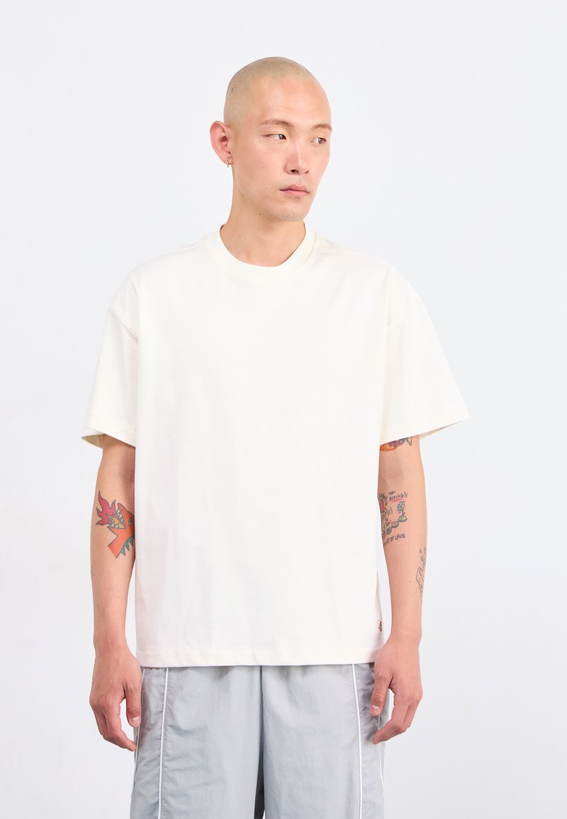 T-shirt a maniche corte color crema con una vestibilità rilassata, scollo a girocollo e una texture di cotone morbida. Nessun motivo o decorazione visibile.