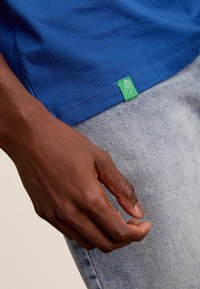 Blauwe katoenen t-shirt met een groen logo-label aan de zoom, gecombineerd met lichtdenim broeken. Close-up opname die zich richt op textuur en kleur.