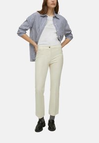 Camicia a righe con bottoni in blu e bianco sopra una t-shirt bianca, abbinata a pantaloni color crema e mocassini neri. Texture sottile e vestibilità classica.