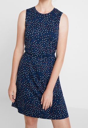 Ärmelloses marineblaues Kleid mit kleinen verstreuten rosa, weißen und blauen Punkten, getragen von einer Person, die vor einem schlichten Hintergrund steht.