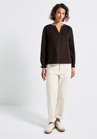 Vrouw staat gekleed in een donkere lange mouw shirt, lichtgekleurde opgerolde broek en beige sneakers tegen een effen witte achtergrond.