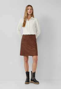 Biała koszula z długim rękawem z guzikami, brązowa zamszowa spódnica oraz czarne botki z grubą podeszwą i teksturowanymi bocznymi panelami.