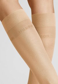 Beige Oberschenkelstrümpfe mit einer glatten Textur, die oben einen verstärkten Bund mit dezent aufgedrucktem Markennamen aufweisen.