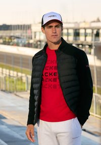 Chaqueta acolchada negra, camiseta roja con estampado de logo, pantalones de chándal grises y una gorra blanca y negra. Diseño elegante con colores en contraste.