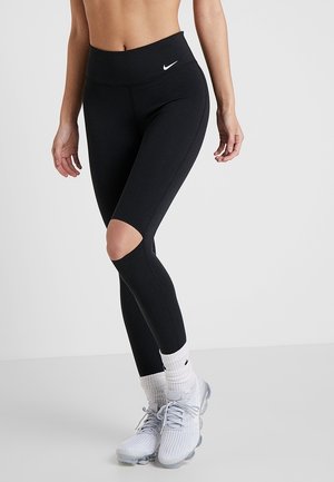 Czarne legginsy sportowe z wycięciem w kolanach, z wysokim stanem i elastycznym materiałem. Połączone z białymi skarpetkami i szarymi butami sportowymi.