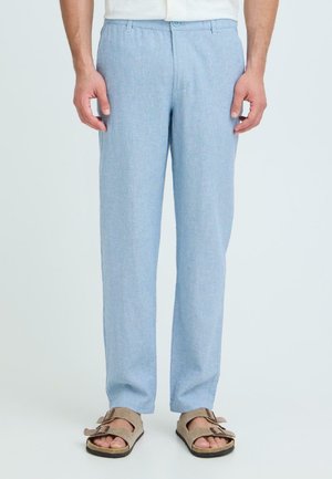 Mand iført lyseblå bukser og beige sandaler med dobbelte remme, stående mod en ensartet lys baggrund.