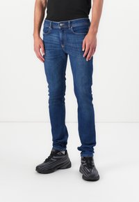Vaqueros de denim azul con un corte ajustado, diseño de cinco bolsillos y costura en contraste. Llevados con zapatillas deportivas negras y una camiseta negra ajustada.