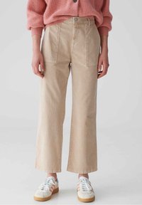 Pantalon beige à jambes larges en tissu coton, doté de poches avant et d'une fermeture à bouton, associé à un pull en maille rose et des baskets blanches.