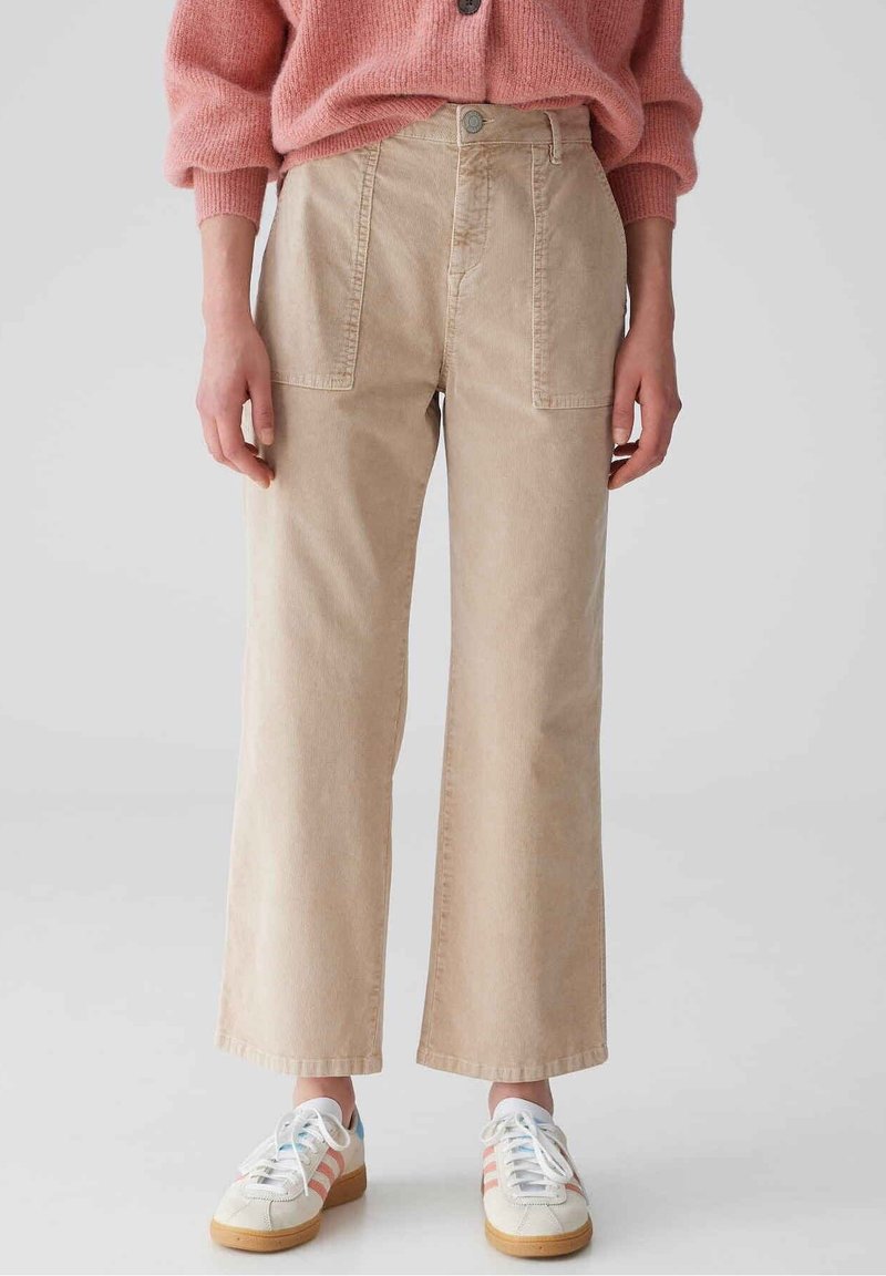 Pantalon beige à jambes larges en tissu coton, doté de poches avant et d'une fermeture à bouton, associé à un pull en maille rose et des baskets blanches.